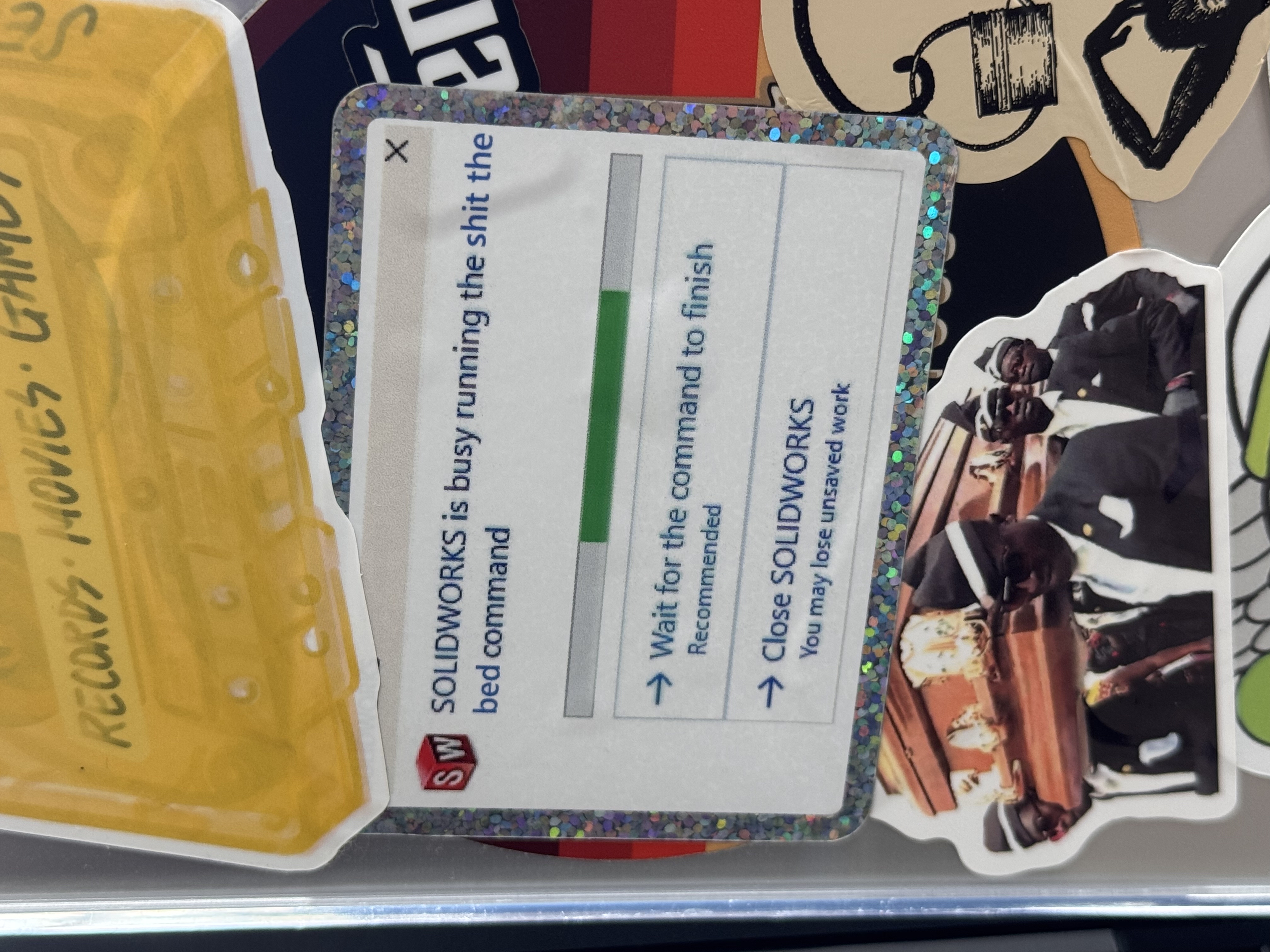 A close-up photo of a MacBook Pro laptop case, with a sticker of a SOLIDWORKS error window edited to say 'SOLIDWORKS is busy running the shit the bed command'.